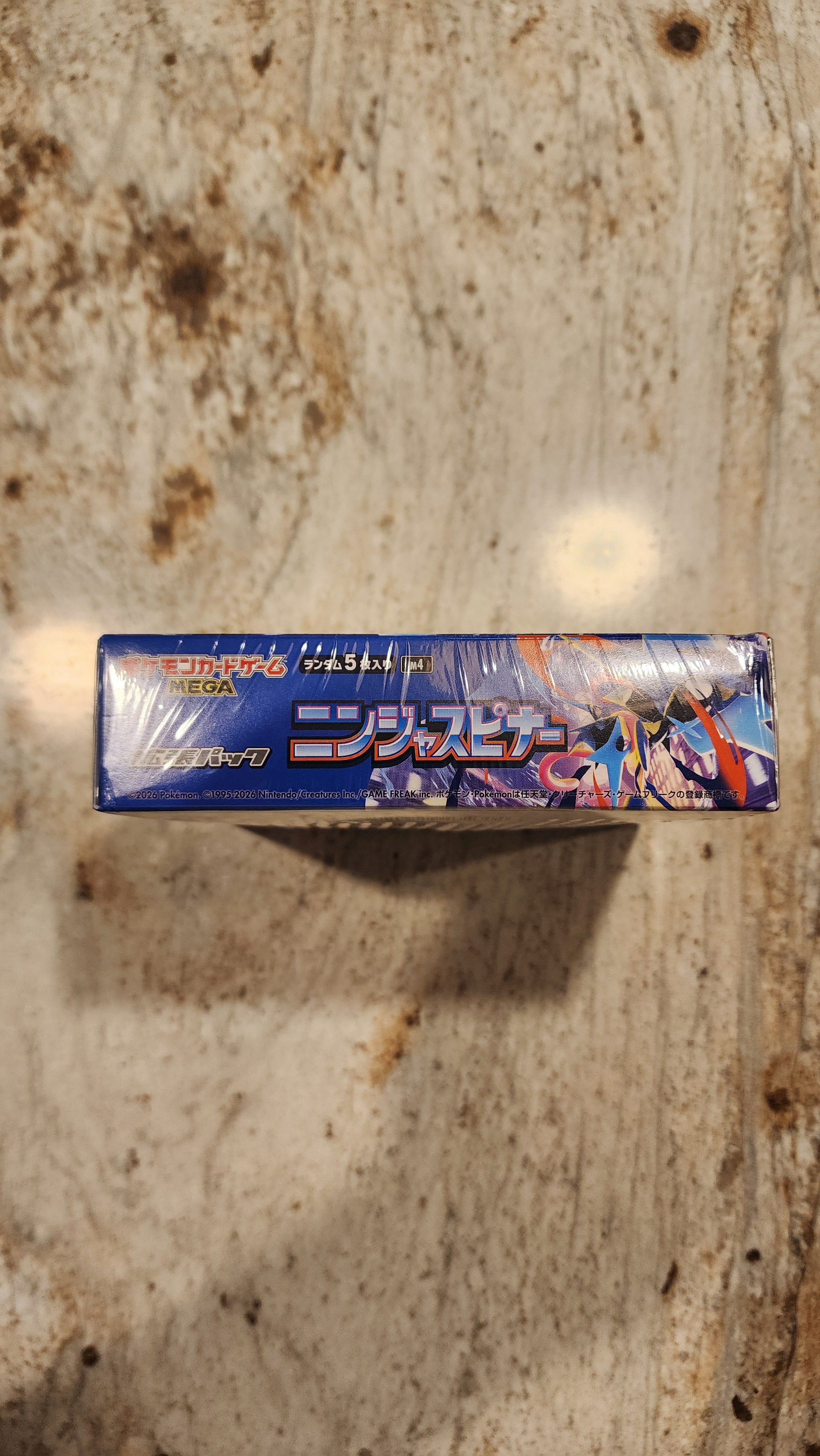 A sealed blue box of Pokémon trading card booster packs standing upright on a worn beige stone surface.
