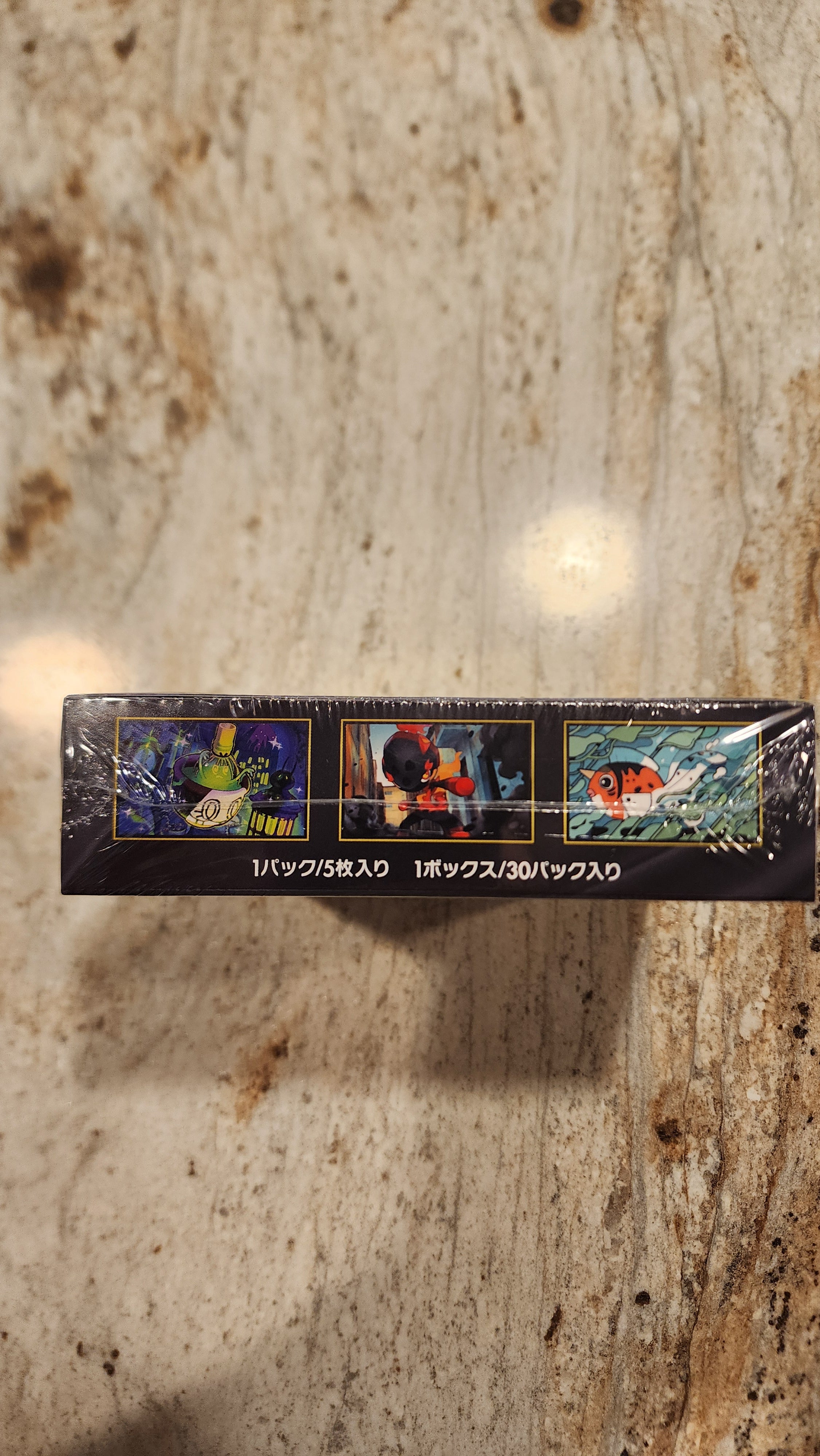 A tall, narrow black product box wrapped in clear plastic, standing vertically on a beige and brown marbled countertop. The box features three small illustrated panels with colorful scenes and vertical white text on the right side.