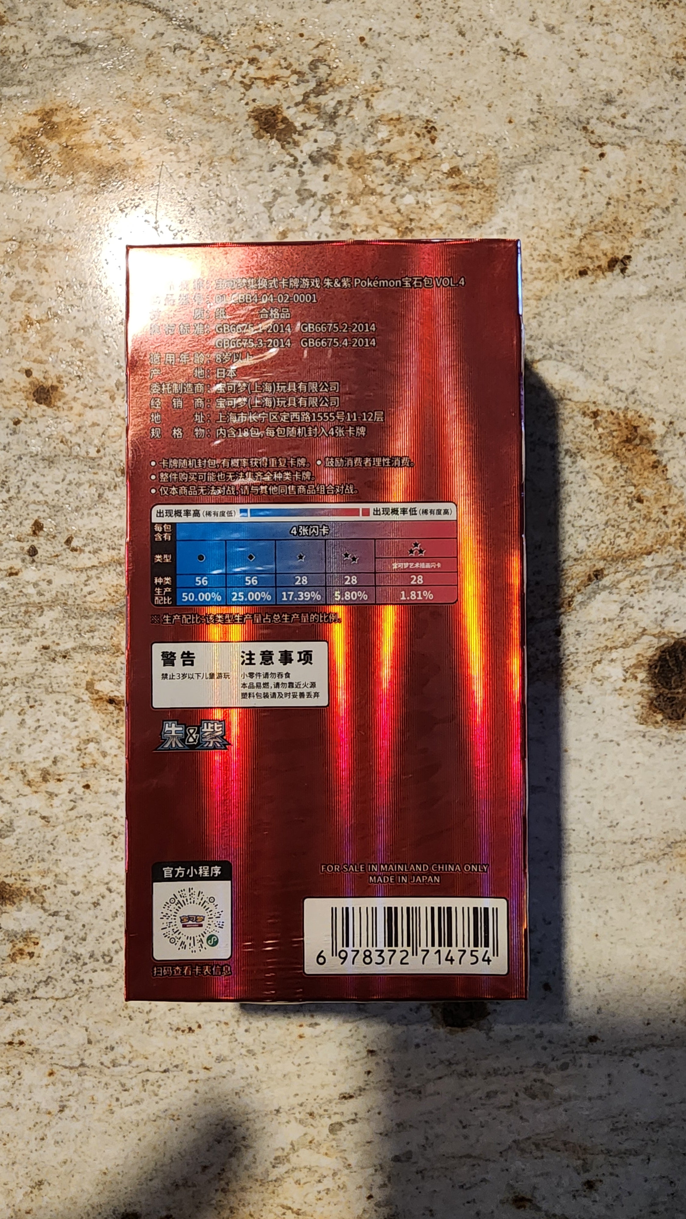The back of a red metallic box with Chinese text, a blue and red warning label, and a barcode, placed on a speckled beige stone surface.