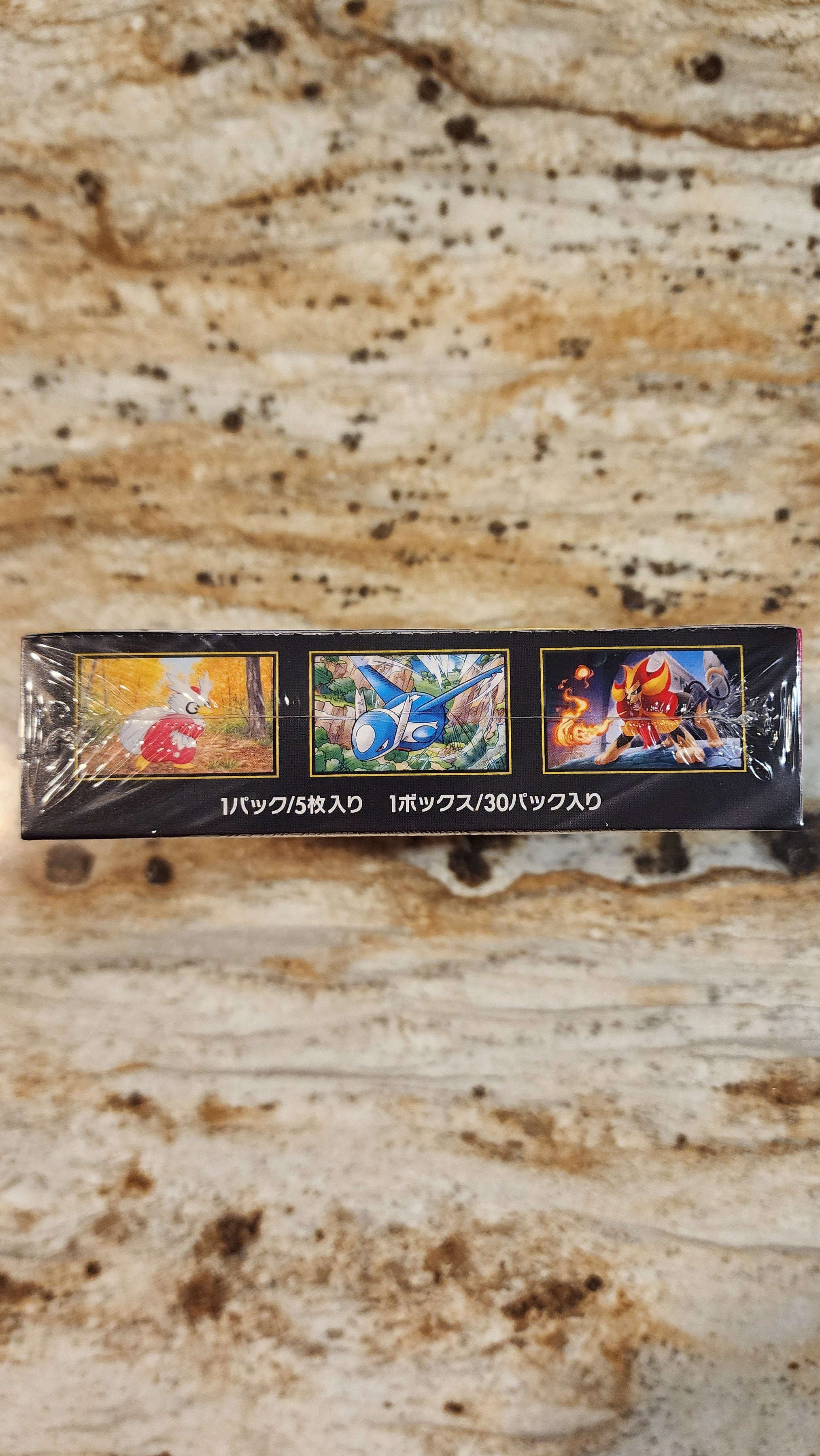 A sealed black box containing three colorful illustrated cards or inserts, photographed on a beige stone surface. The box has Japanese text along the bottom and small fantasy/game-style artwork on each panel.