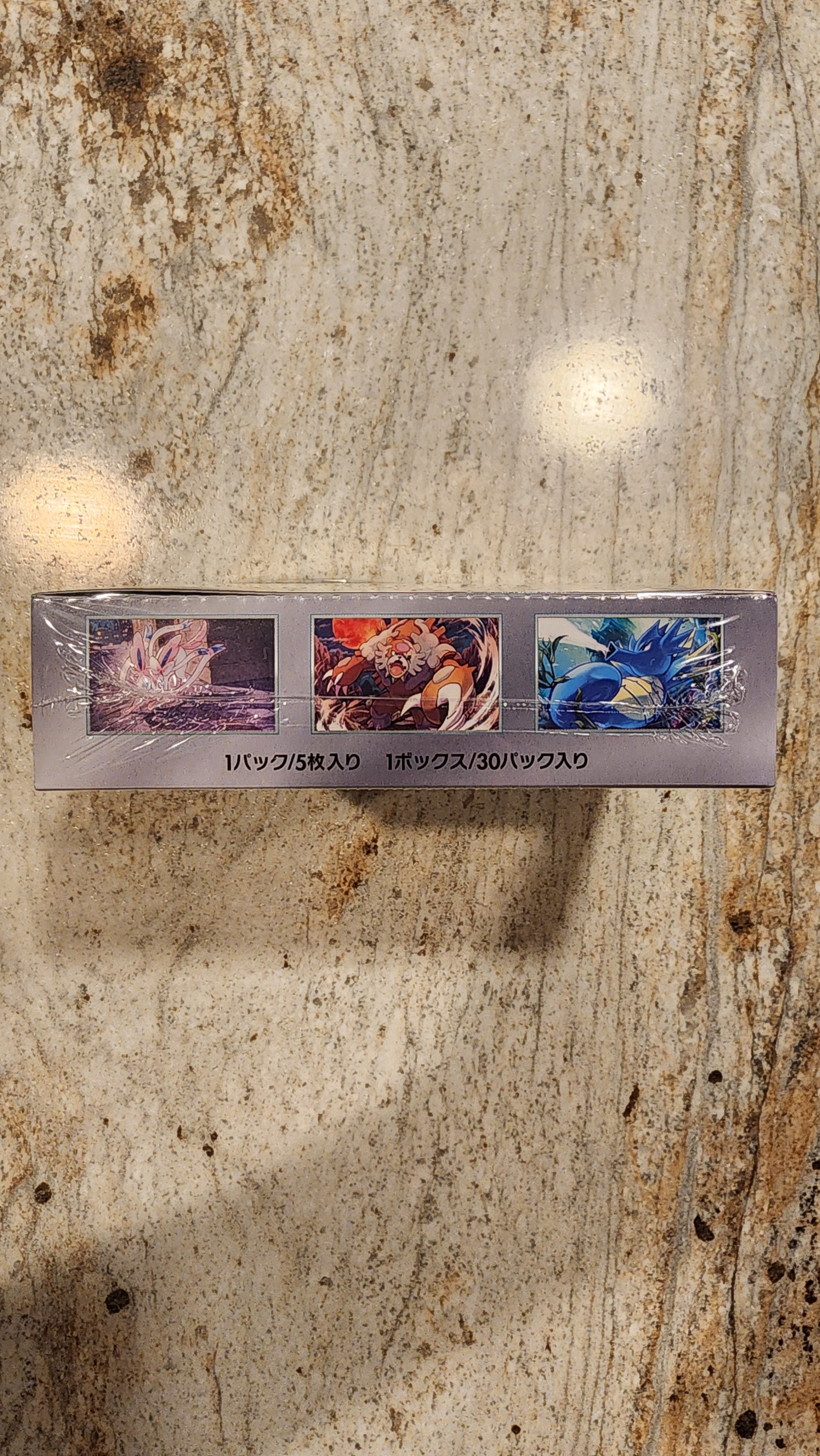 A slim rectangular product box wrapped in clear plastic, standing upright on a mottled beige stone surface. The box is light gray with three vertical panels showing colorful abstract artwork and small black text running down the center.