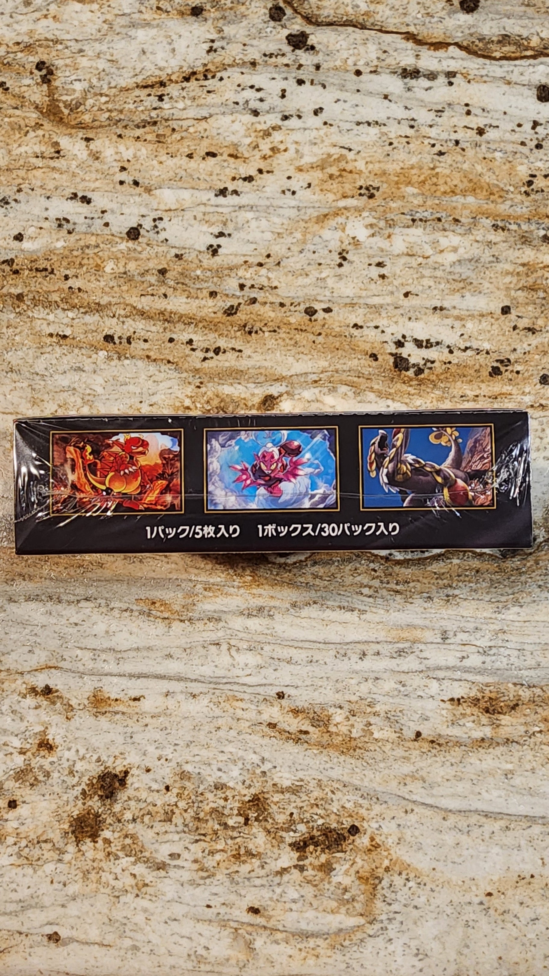 A small rectangular black product box with three colorful printed illustrations sits on a beige stone surface. The box appears to be the item being sold, photographed from above.