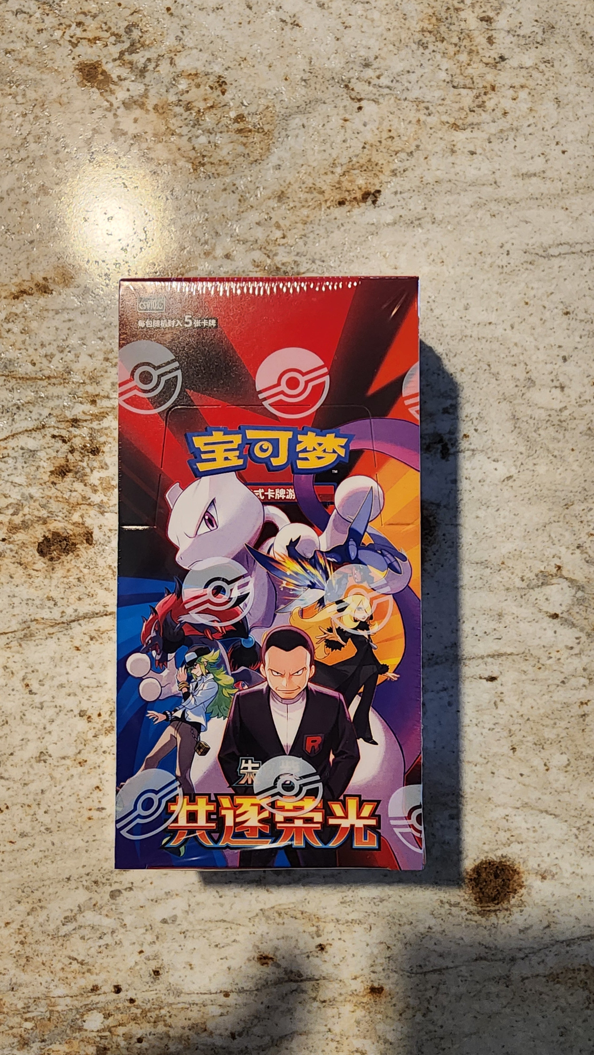 A Pokémon booster pack box featuring colorful character artwork, photographed standing on a mottled stone-like surface.