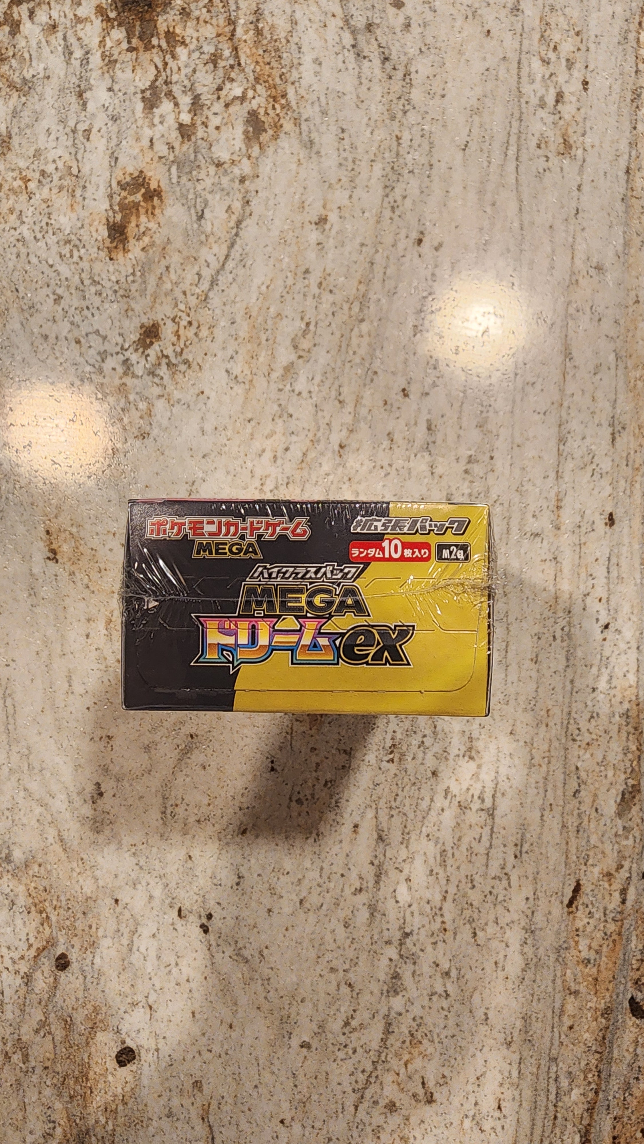 A sealed box of Pokémon Booster Packs, featuring a yellow and black design, placed on a textured stone surface.