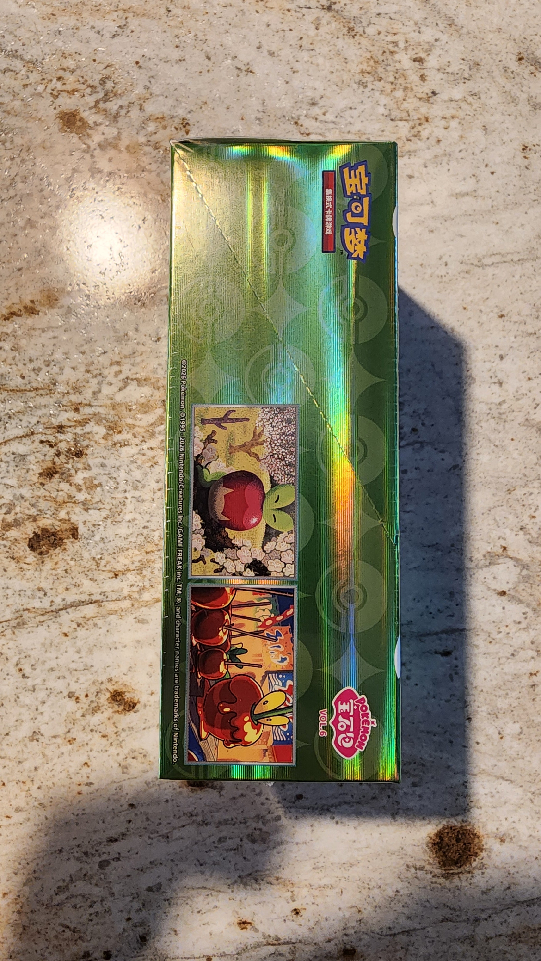 A green holographic Pokémon trading card box standing upright on a speckled beige stone surface, with bright sunlight casting a strong shadow to the right.