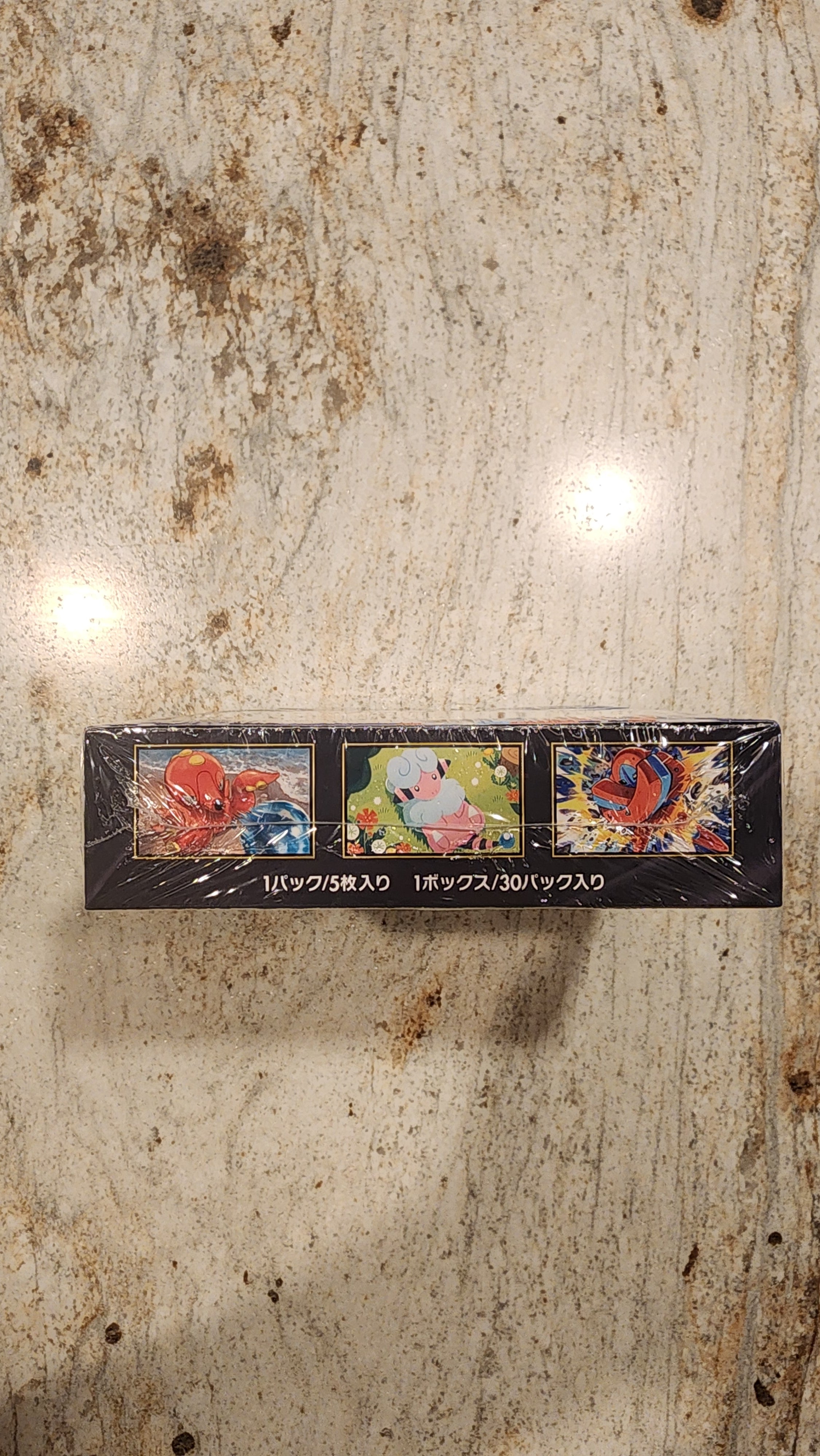 A sealed black rectangular box with three colorful illustrated panels and white vertical text, photographed from above on a mottled beige stone-like surface.