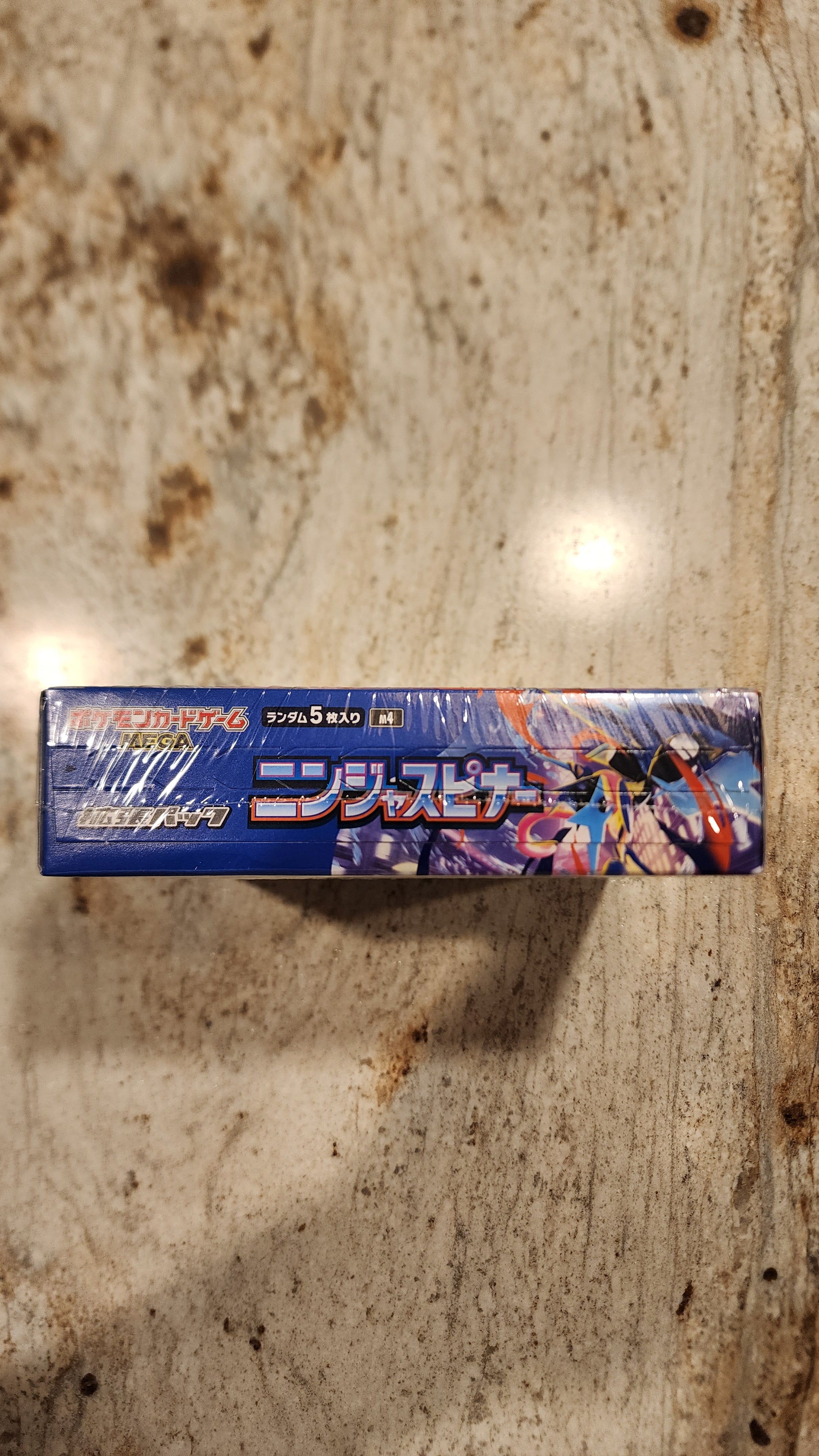 A boxed product standing upright on a mottled beige stone surface, with Japanese text and colorful anime-style artwork on the front and side panels.