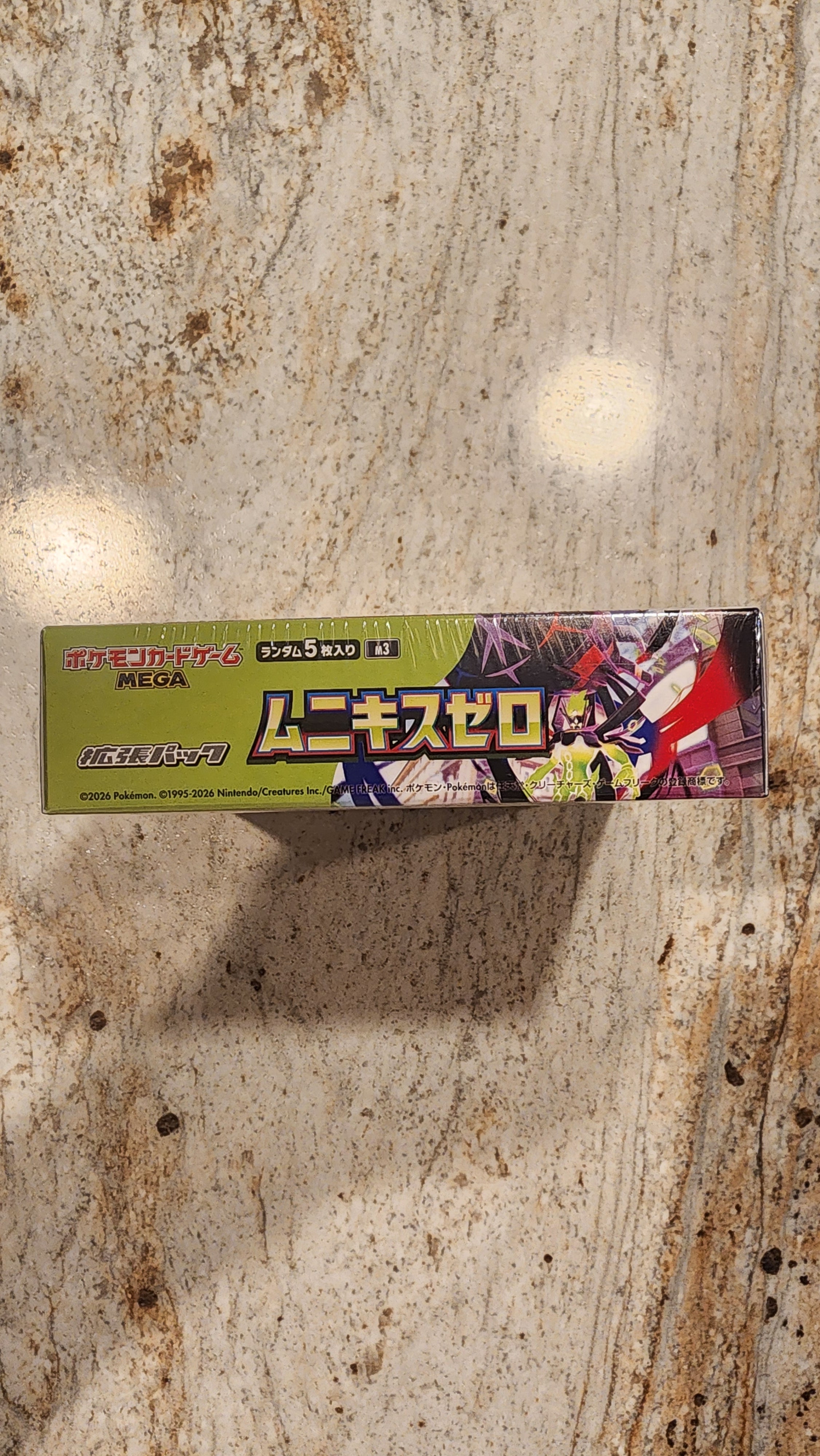 A long, narrow product box with Japanese text and colorful anime-style artwork, standing upright on a mottled beige stone surface.