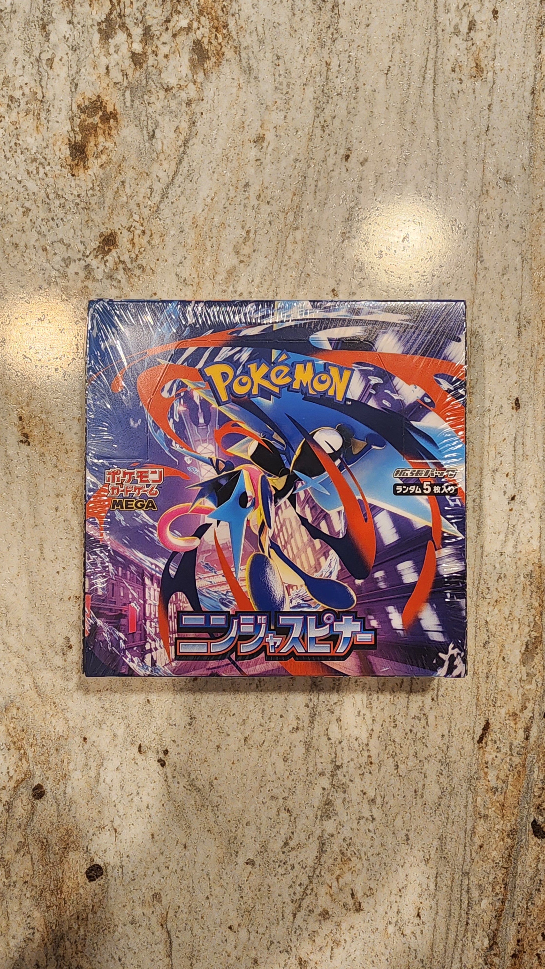 A sealed Pokémon trading card booster pack from the Monstera set, featuring a dynamic blue and orange character illustration, placed on a textured beige stone surface.