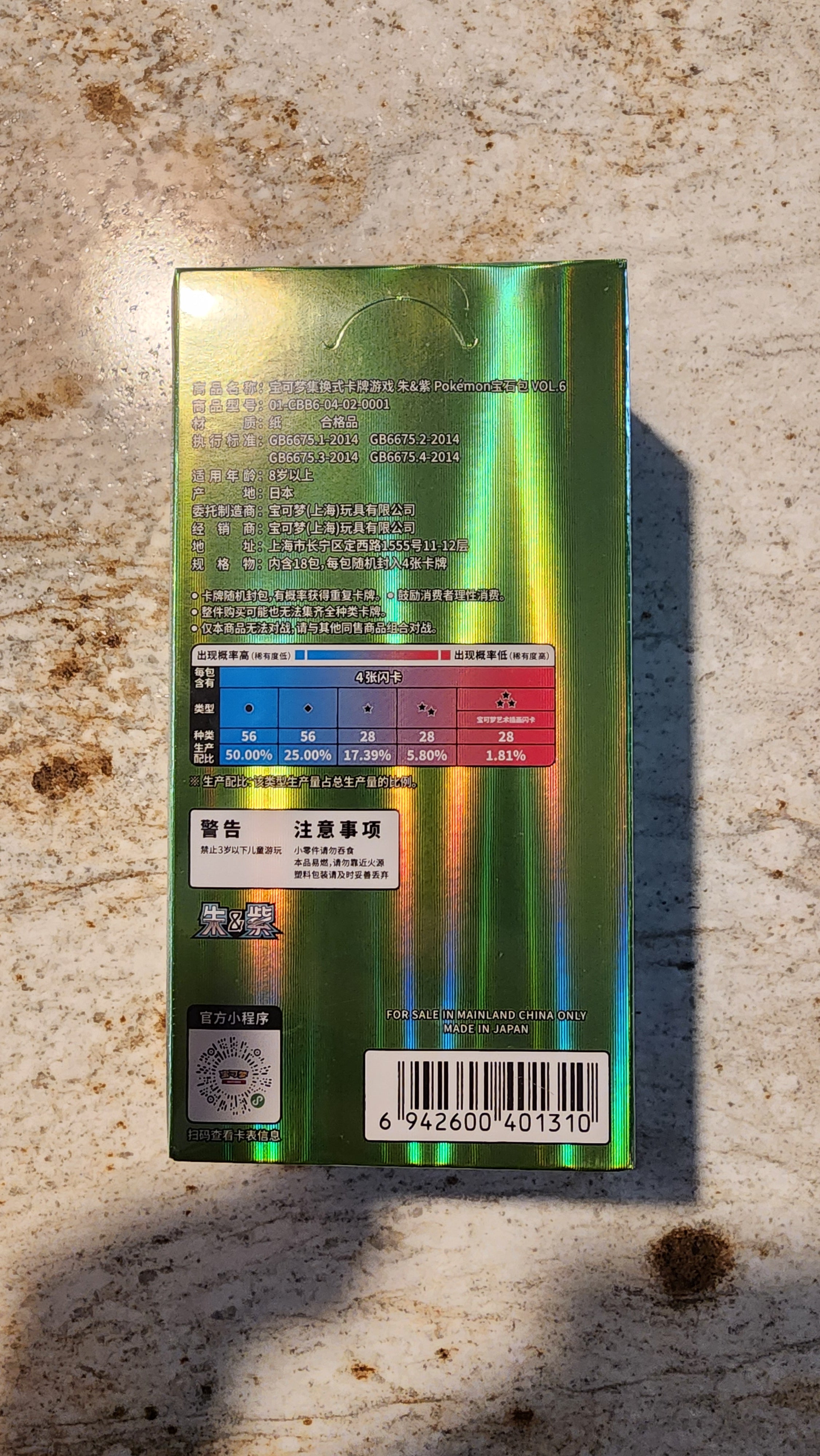 The back of a green holographic box with Chinese text, a color chart, and a barcode, lying on a textured beige stone surface.