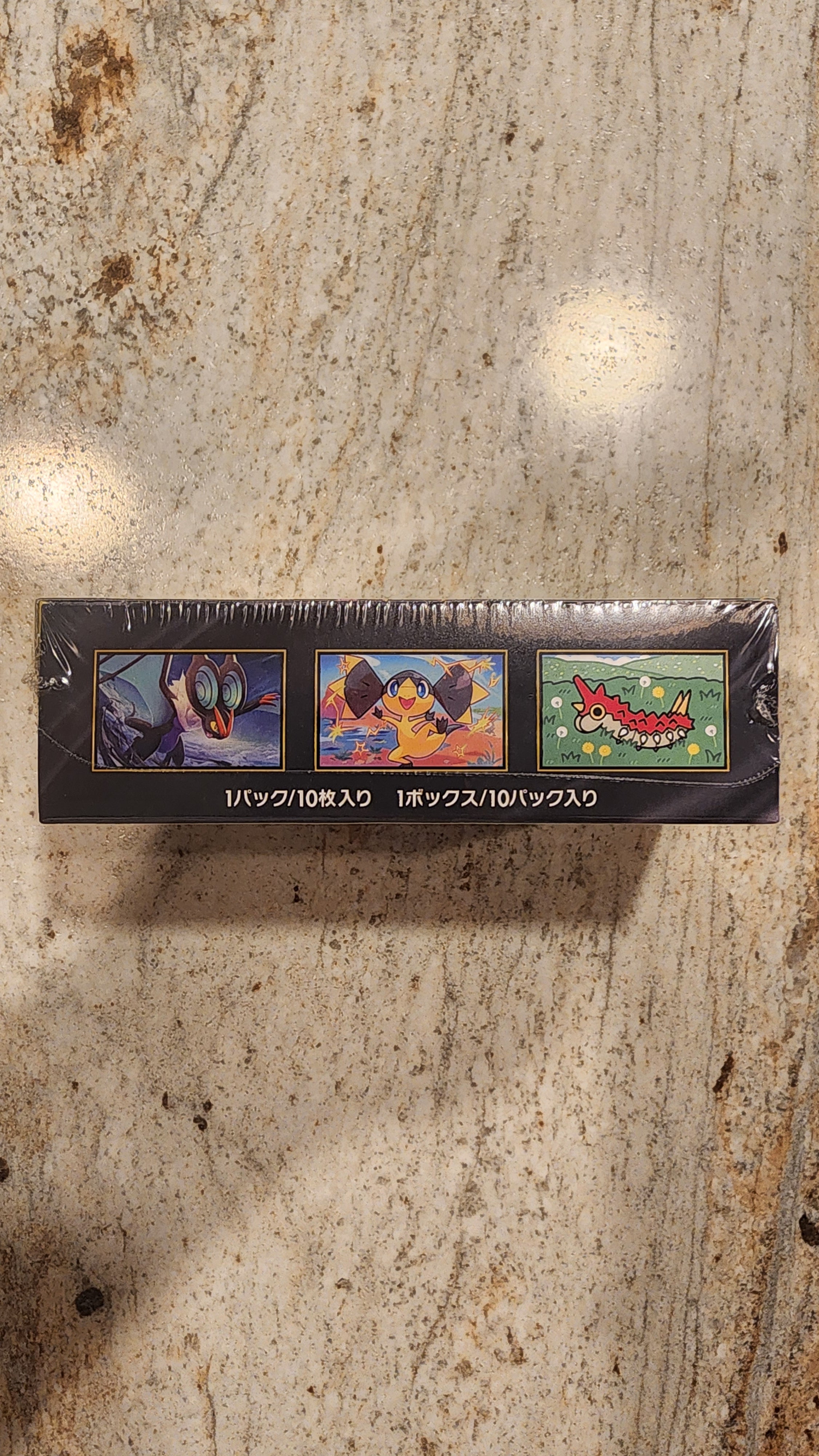 A tall, narrow black product box wrapped in clear plastic, standing upright on a beige stone surface. The box features three colorful illustrated panels and vertical Japanese text.