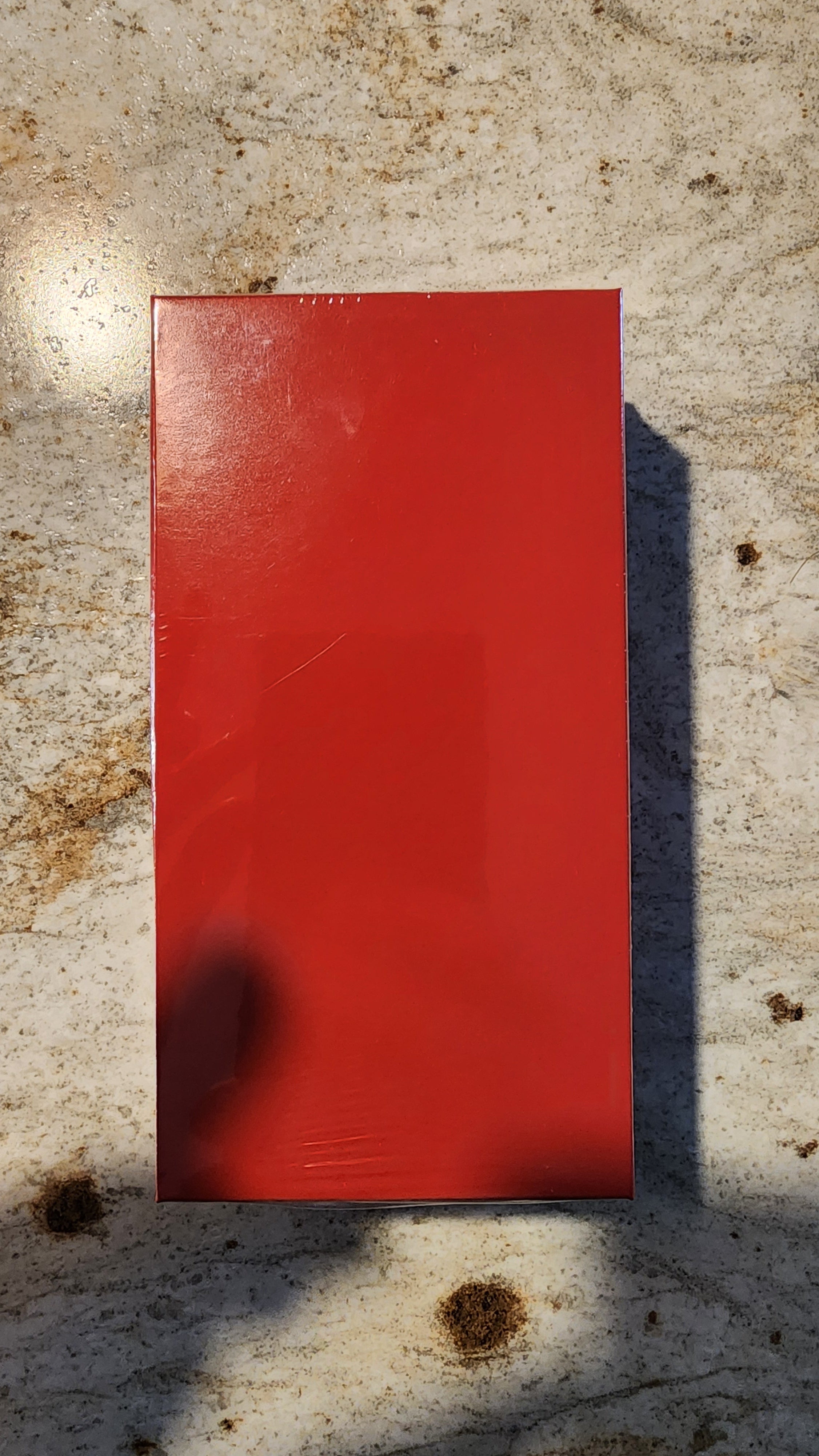 A solid red rectangular box or package lying on a textured concrete surface with strong sunlight and shadows.