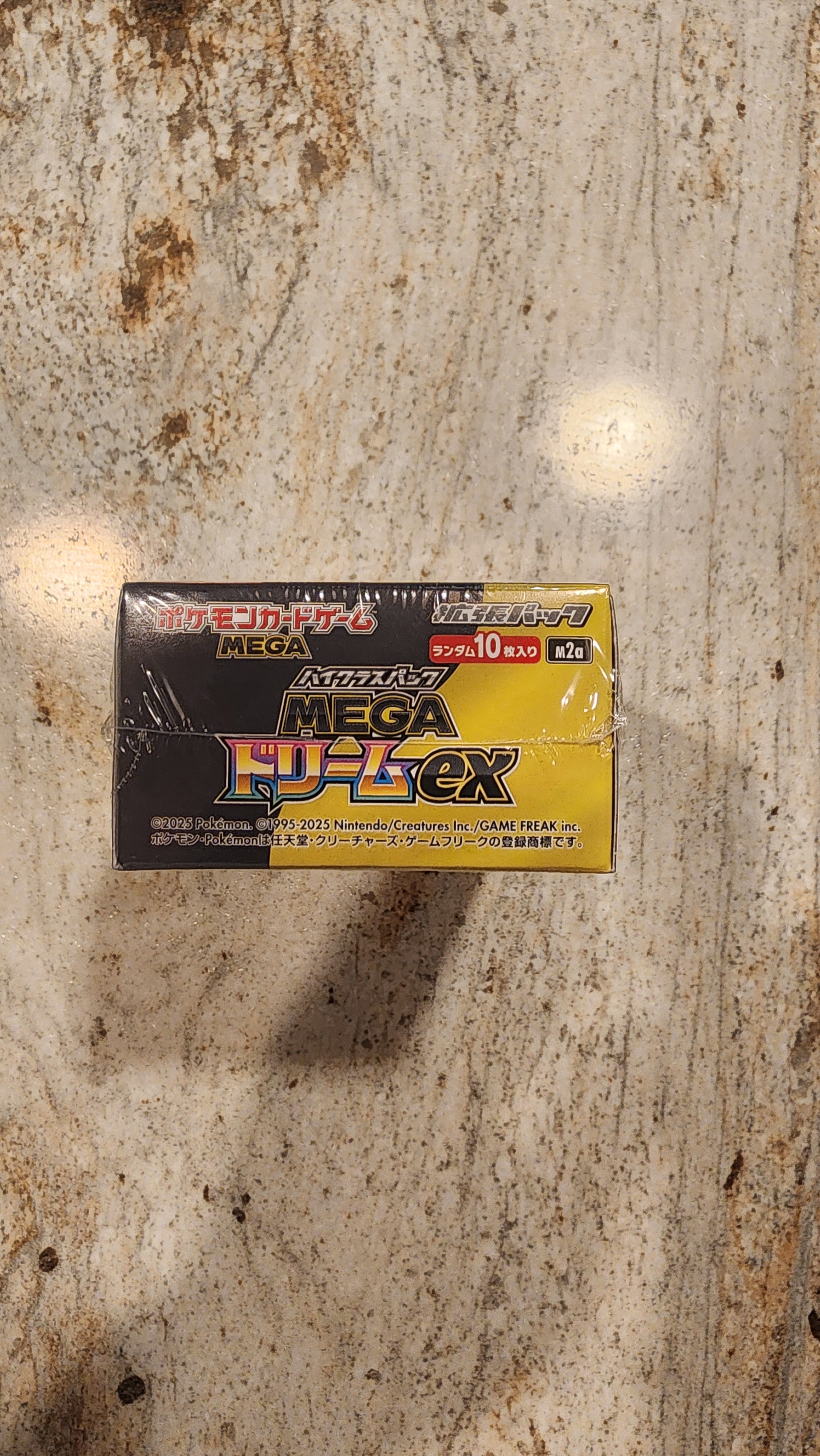 A sealed yellow and black box of Pokémon cards sits on a textured beige stone surface.