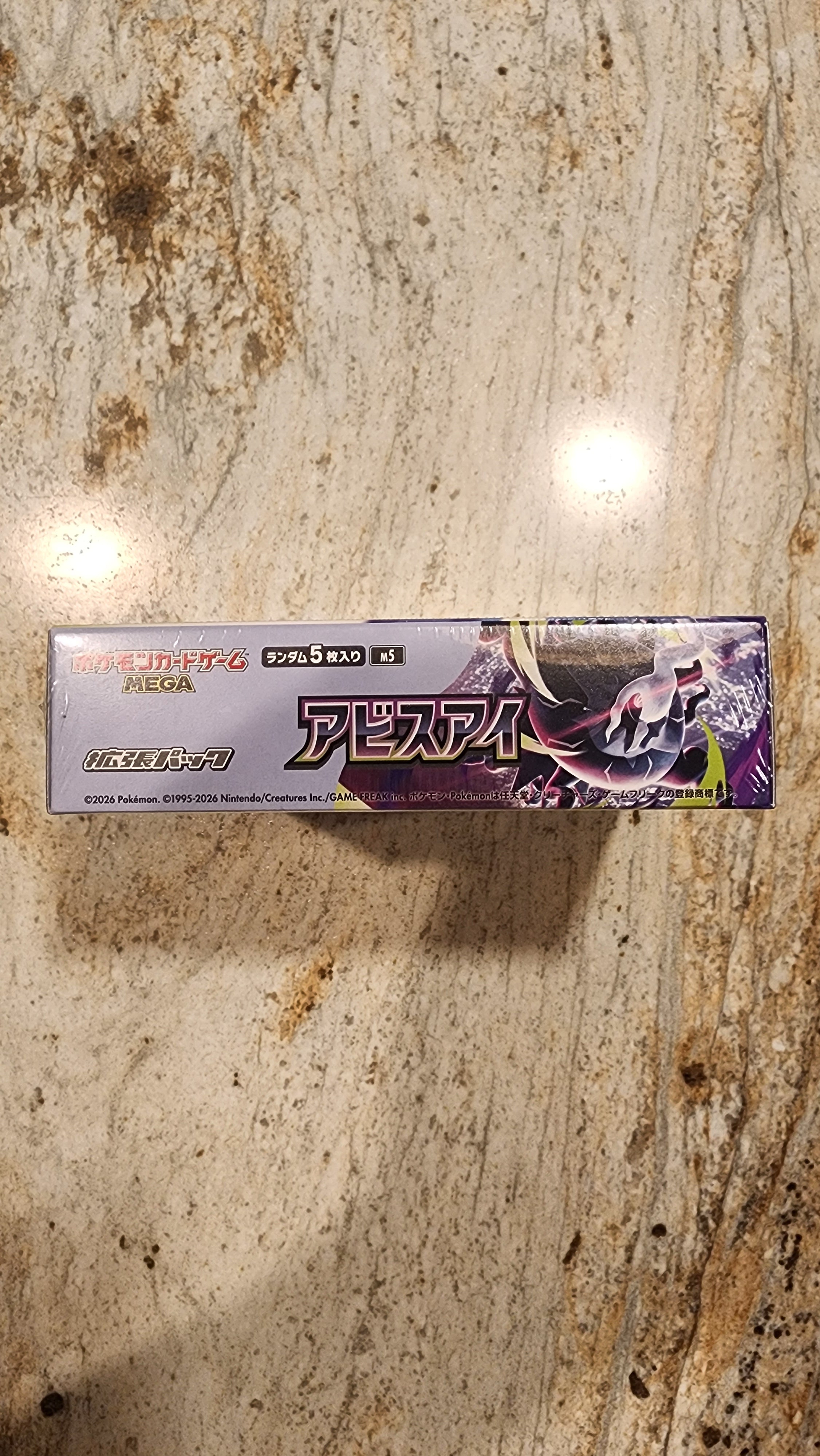 A boxed product with Japanese text and anime-style artwork, standing upright on a speckled beige stone surface.