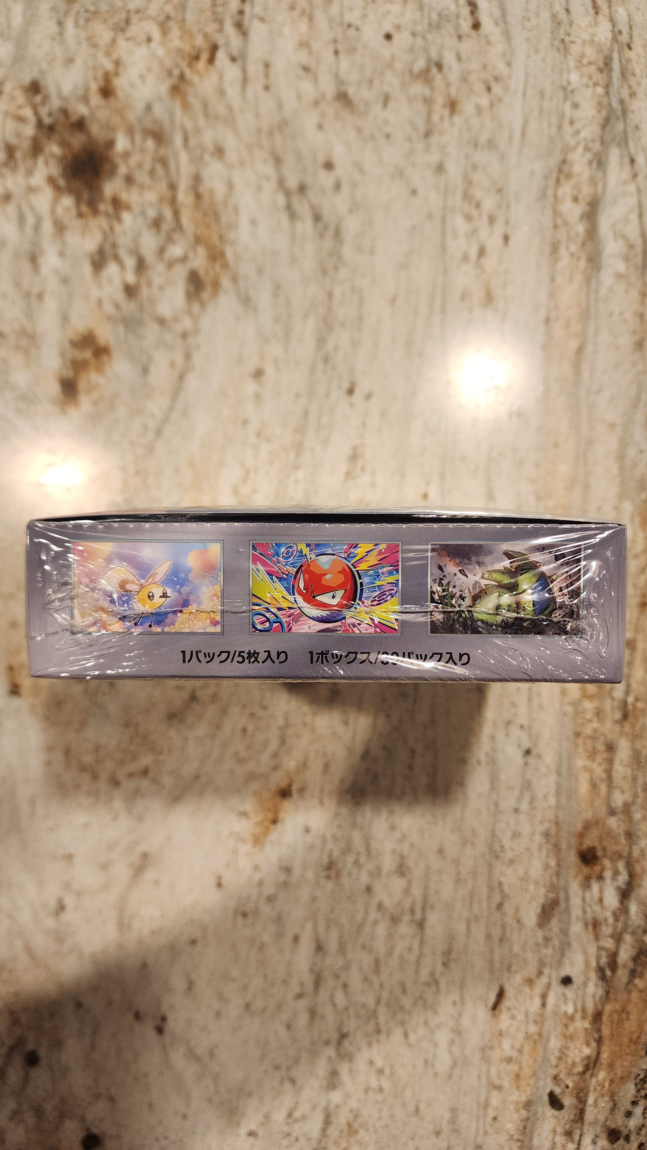 A shrink-wrapped Pokémon TCG card box standing upright on a speckled beige stone surface. The box is mostly dark gray/black with three colorful illustrated card panels visible on the front and vertical text including 'OVERGARUM' and 'POKEMON'.