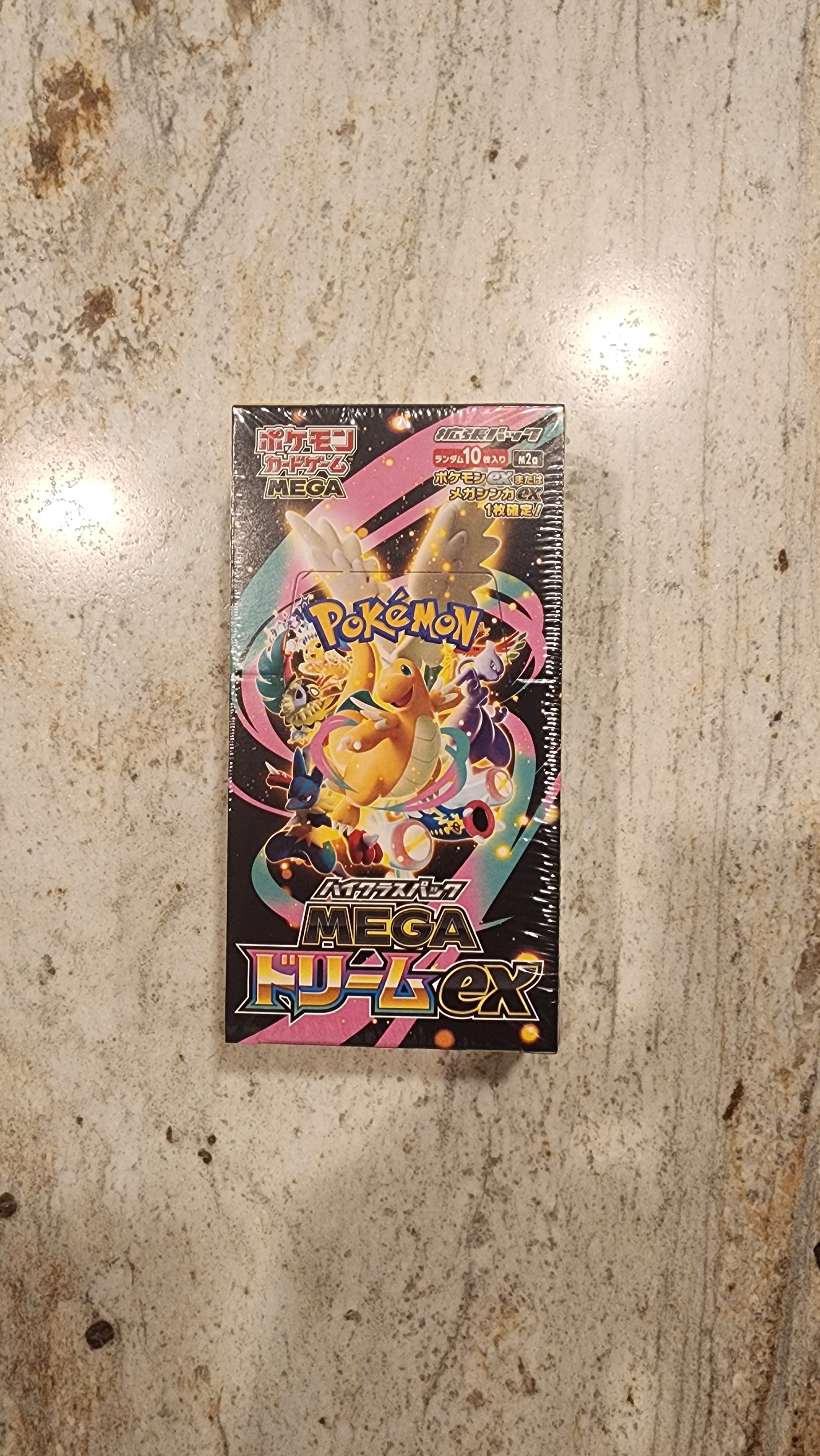 A sealed Pokémon TCG Mega booster box featuring colorful character artwork, placed on a light beige stone or laminate surface.