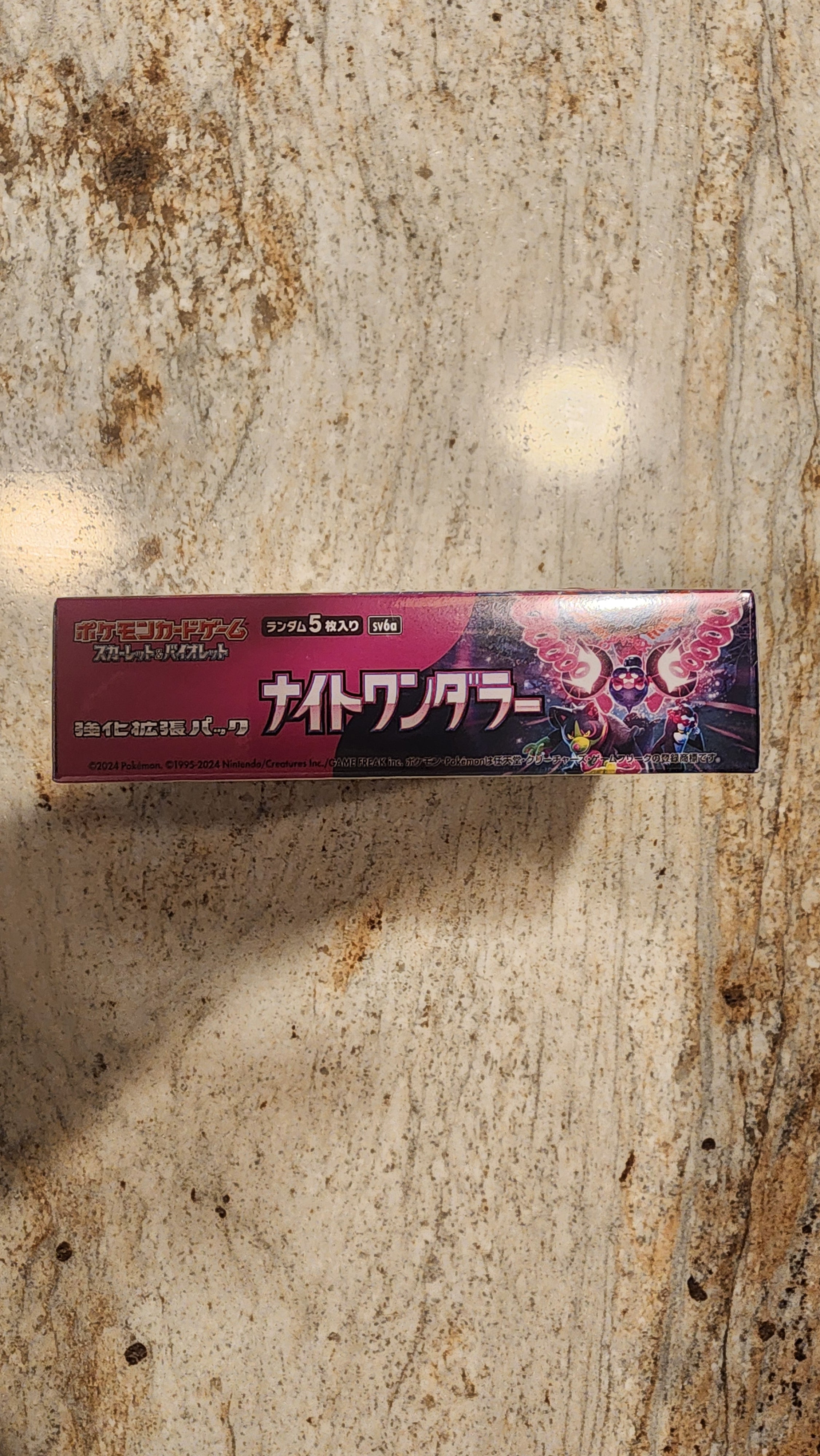 A tall rectangular product box with Japanese text and a dark pink/red design, photographed standing upright on a mottled beige stone-like surface.