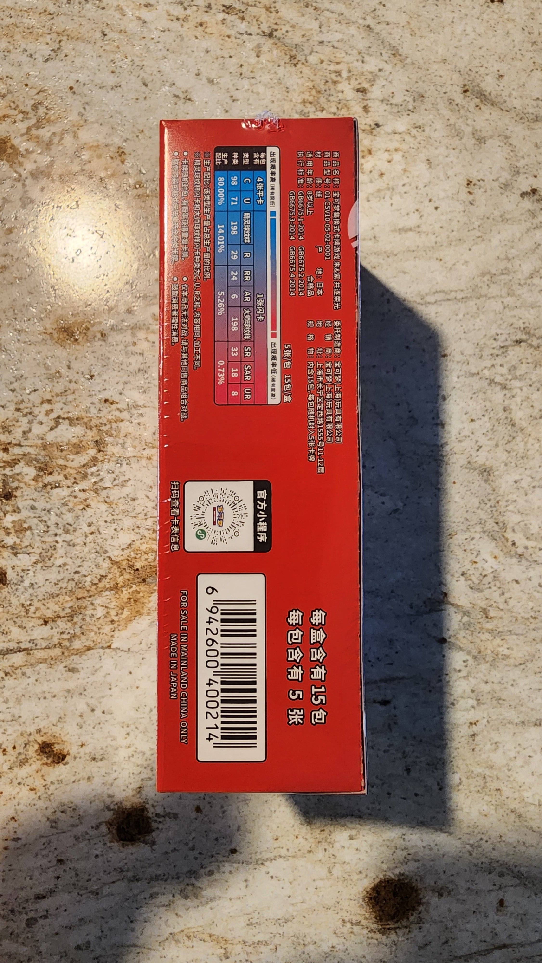 A red rectangular product box photographed from above on a mottled beige stone-like surface. The box has printed text, a barcode, and a small multicolor ingredient or nutrition-style panel near the top.