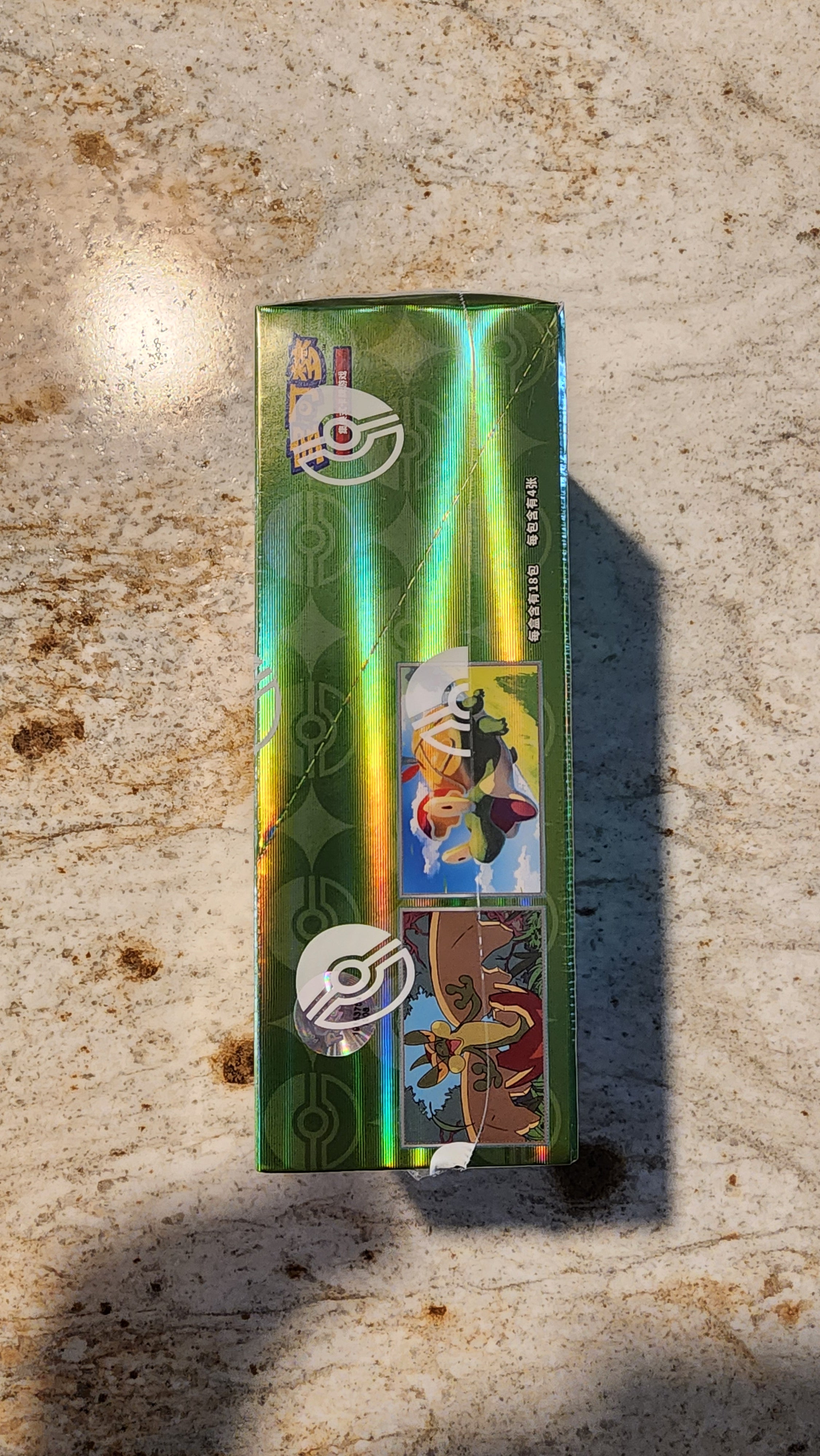 A long, narrow green holographic box or package standing upright on a textured beige stone-like surface. The package has colorful printed artwork, a Pokéball logo, and vertical text reading 'SERPENTIN KEBUTUAN'.
