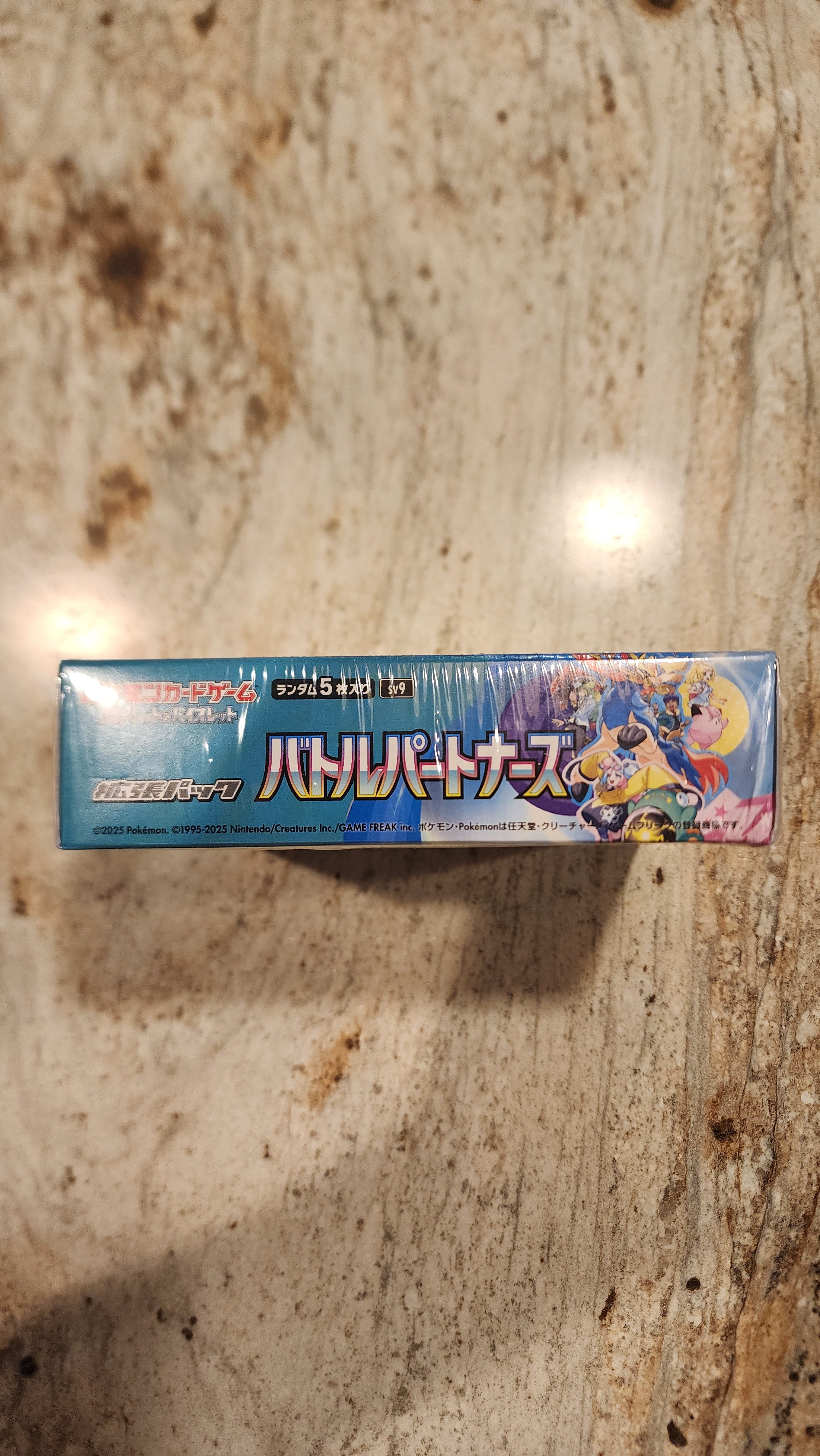 A boxed product with colorful anime-style artwork standing upright on a mottled beige stone surface. The package is predominantly blue with white and pink text and graphics.
